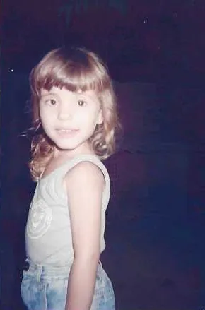 Raíza ainda criança, sorridente, cabelo loiro cacheado, vestindo regata e jardineira jeans, em ambiente pouco iluminado.