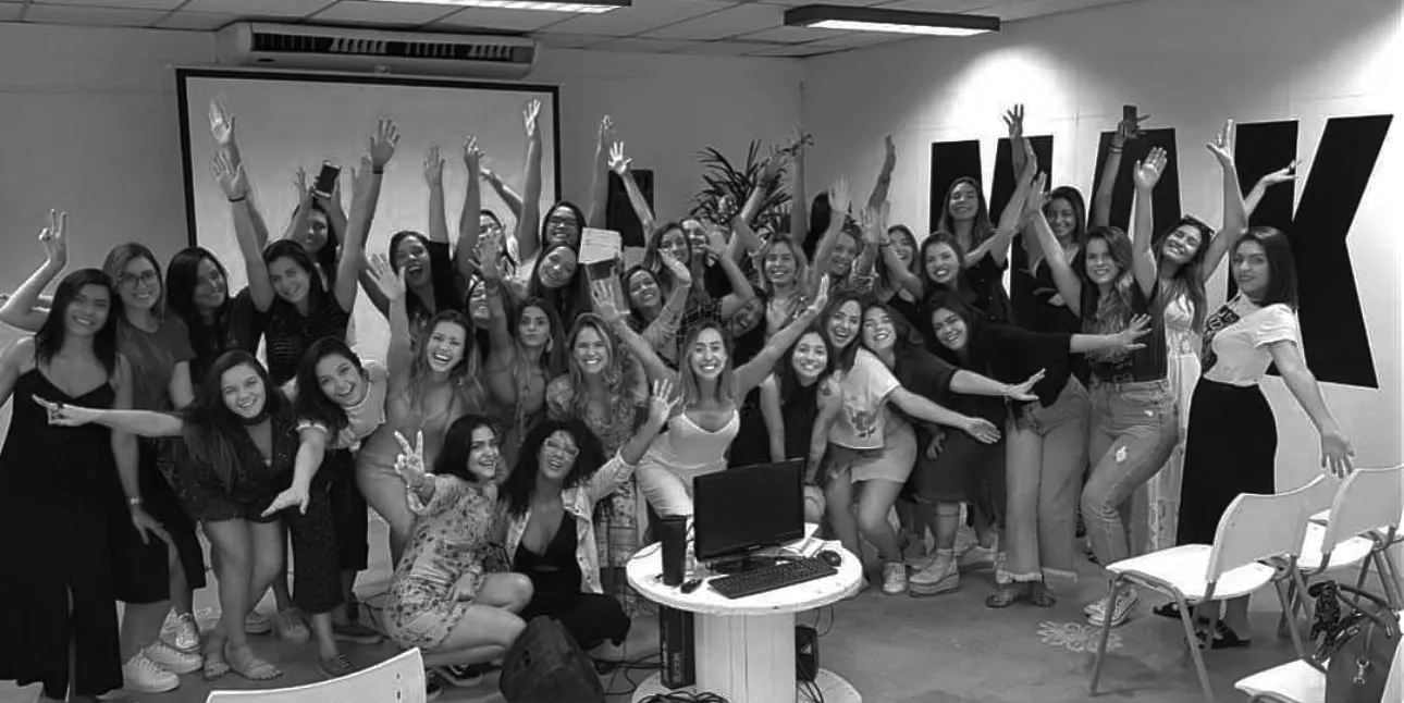 Grupo de mulheres comemorando com os braços erguidos em sala de treinamento, com telão e letreiro ao fundo.